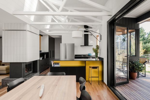 Bright open-plan kitchen and dining area with a light timber dining table, black cabinetry, and a vivid yellow kitchen island. White exposed roof trusses and a central fireplace define the space, while large black-framed sliding glass doors open to a timber deck and planted outdoor area.
