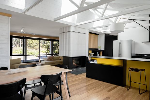 Open-plan contemporary kitchen, dining and living area with a bright white exposed-truss ceiling and skylights. The kitchen features black cabinetry, a vivid yellow island accent, stainless-steel appliances and timber flooring. A dining table and black chairs sit in the foreground, while a lounge area and extensive black-framed glazing are visible beyond. A central fireplace is enclosed by a textured white feature wall.