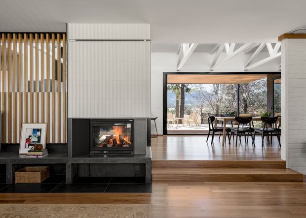 Bright contemporary open-plan living and dining area centred on a black built-in fireplace. The living room features warm timber flooring, a dark stone or tiled hearth and low built-in storage, with a vertical timber slat screen and white ribbed wall treatment beside the fireplace. A step leads into the dining area, which has a timber table and black chairs beneath exposed white roof trusses. Large glazed doors open to an outdoor terrace and wooded setting.