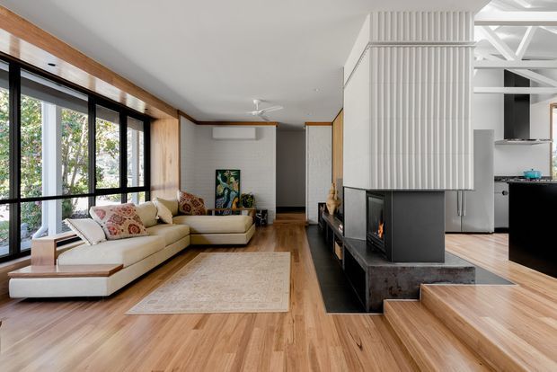 A bright, contemporary open-plan living room with a large cream sectional sofa arranged beside floor-to-ceiling black-framed glazing. Warm timber flooring and ceiling beams contrast with white walls and a sculptural black fireplace set into a dark stone hearth. A white vertically ribbed partition separates the living area from a partially visible kitchen, while timber steps lead to the adjacent level.