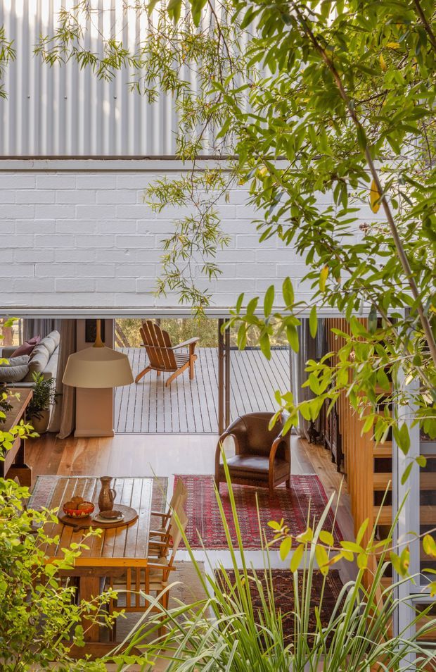 A warm, plant-filled interior courtyard or living space viewed through dense greenery. The room features white-painted brick walls, corrugated metal cladding above, timber flooring, exposed timber-framed glazing or sliding doors, a rustic timber dining table, leather armchair, patterned rugs, and a broad pendant lamp. An outdoor deck with timber seating is visible beyond the open glazing, creating a strong indoor-outdoor connection.