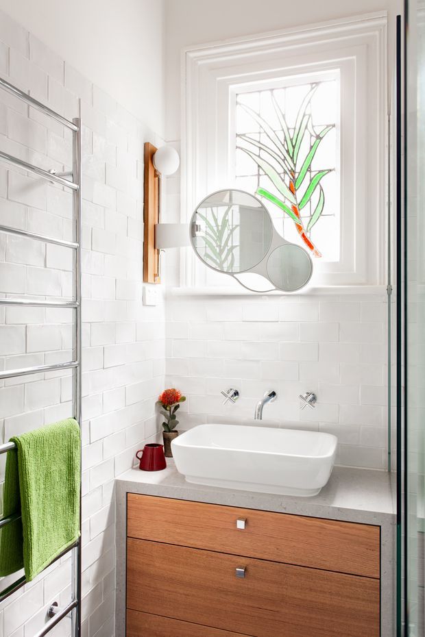 Bright, compact contemporary bathroom with white subway-tile walls, a wall-mounted timber vanity with grey stone benchtop, white vessel basin, chrome tapware, heated towel rail, frameless glass shower enclosure, and a decorative stained-glass window. A round multi-panel mirror and small decorative accents add character.