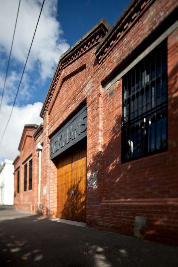 Low-angle exterior photograph of a historic red-brick industrial building in an urban laneway. The facade features decorative brick detailing, tall black-framed barred windows, and a large timber loading door beneath a dark sign reading “CANAL”. Strong sunlight and deep shadows emphasize the masonry texture and heritage character.
