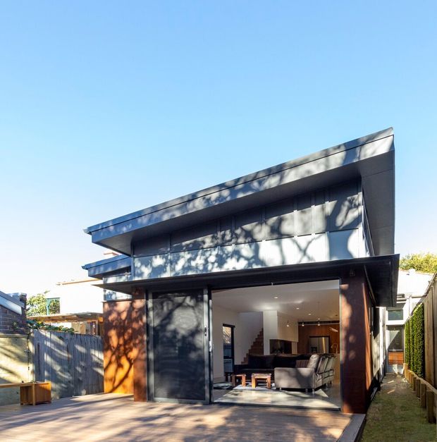 Exterior view of a contemporary two-storey residential home with a strong horizontal, flat-roofed form. Dark cladding and timber-toned elements frame a large open glazed garage or living opening, with a visible open-plan interior and staircase. The house sits on a narrow suburban site with a paved forecourt and side garden.