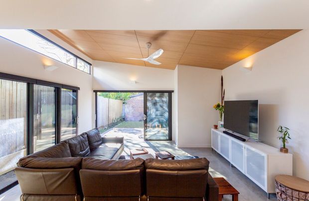 Bright contemporary living room with a large brown leather sectional sofa facing a wall-mounted television and low white media console. The room has expansive sliding glass doors opening to a small courtyard, high clerestory glazing, a warm timber-lined ceiling with a ceiling fan, white walls, and polished light flooring.