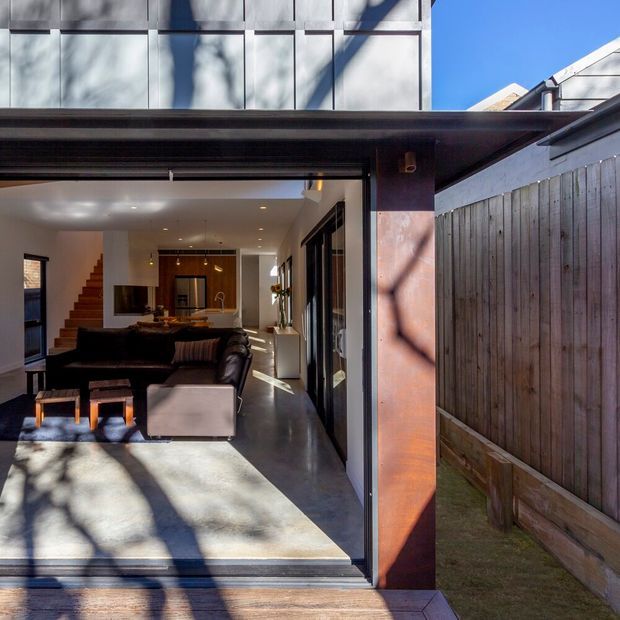 View from an outdoor patio into a contemporary open-plan living room through fully opened glazed doors. The interior features a dark sectional sofa, timber staircase, polished concrete flooring, recessed lighting, and warm timber joinery. The exterior shows a timber privacy fence, a covered roof edge, and a modern upper facade with large panelled glazing or cladding. Strong dappled sunlight and shadows create a warm indoor-outdoor connection.