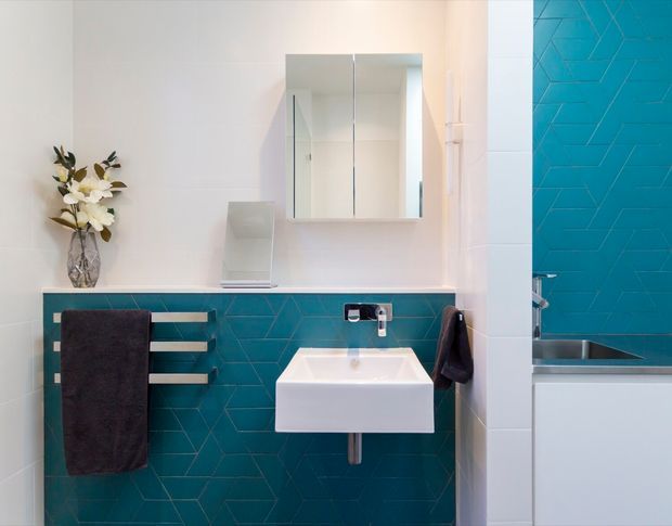 Bright contemporary bathroom featuring a wall-mounted white rectangular basin, mirrored shaving cabinet, chrome tapware, and a teal geometric tiled feature wall. A matching teal tiled bath area is visible through the opening on the right, with dark towel rails and a small decorative vase adding contrast.