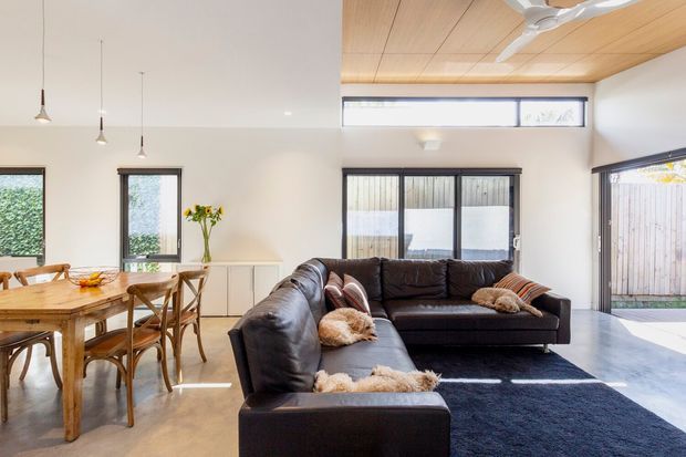 Open-plan living and dining area with a large dark leather sectional sofa, timber dining table and chairs, polished concrete floor, white walls, expansive dark-framed glazing, and a warm timber-lined ceiling with ceiling fan. The bright contemporary room has a relaxed, refined atmosphere and connects visually to an outdoor area through the glazing.