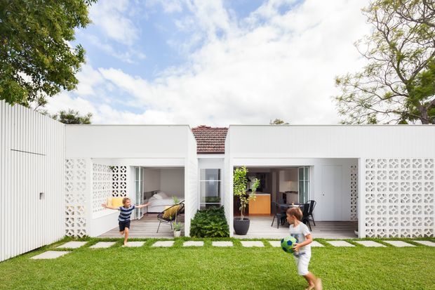Bright exterior view of a low, white residential dwelling opening onto a manicured lawn and paved stepping-stone path. The facade combines vertical white cladding with decorative breeze-block screens, large glazed openings, and a central section with a red tiled pitched roof. Two connected outdoor-facing living areas and interior furnishings are visible through the openings, with mature trees framing the property.