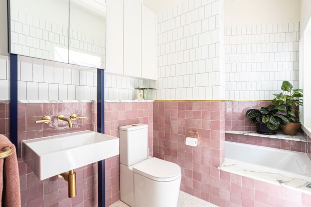 Bright contemporary bathroom with pink glazed wall tiles covering the lower walls and white rectangular tiles above. The room includes a wall-mounted white basin with brass tapware and plumbing, a white toilet, and a built-in bathtub beneath a window ledge with plants. Large mirrored cabinetry and dark blue vertical framing add contrast, while terrazzo-style flooring and brass details contribute a refined, playful character.