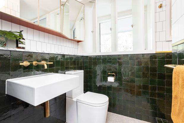 Bright, compact contemporary bathroom with a wall-mounted white rectangular basin, white toilet, brass wall-mounted tapware, and glossy dark green square wall tiles. White upper subway-style tiles, large mirrors, and tall windows create a light-filled space, with a warm wood-toned shelf and mustard towel adding contrast.