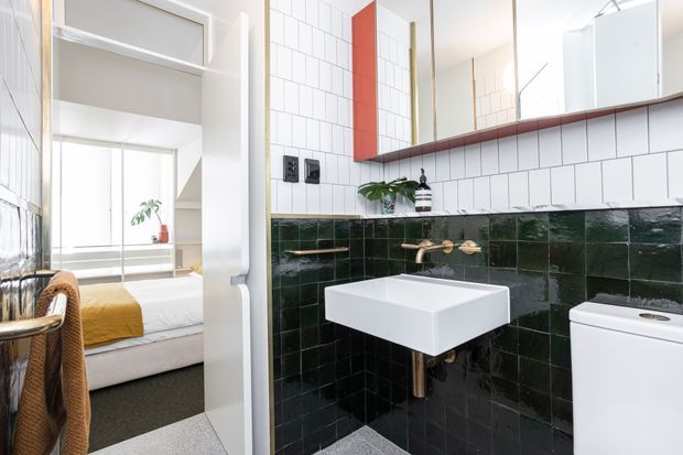 A bright bathroom viewed through an open doorway, featuring a wall-mounted rectangular white basin, brass tapware, white subway wall tiles, dark glossy green lower wall tiles, a white toilet, and a mirrored wall cabinet with timber-toned trim. A bedroom with a bed, mustard throw, and large glazed opening is visible beyond the bathroom.