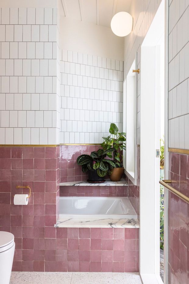 Bright, compact bathroom with a built-in bathtub set beneath a window and marble sill. The walls are clad in glossy white rectangular tiles above a band of glossy dusty-rose/mauve tiles, with terrazzo flooring, brass fittings, white sanitaryware, potted plants, and a spherical pendant light. A partially open white door reveals the exterior beyond.