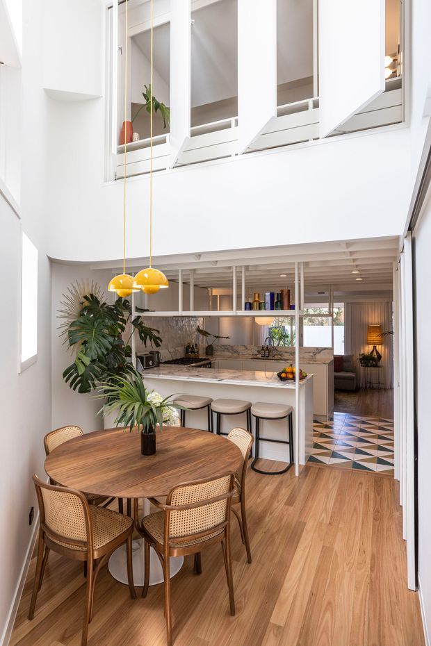 Bright open-plan dining and kitchen interior with a double-height void and upper-level white cabinetry or storage overlooking the space. A round timber dining table with cane-back chairs occupies the foreground, while the kitchen behind features a marble-look island and backsplash, stools, open shelving, and warm pendant lights. Timber flooring, abundant white surfaces, indoor greenery, and a patterned tiled passage create a refined contemporary feel.