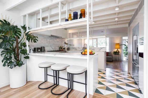 Bright open-plan kitchen with a white island and three stools, marble-look backsplash and benchtops, extensive open shelving, integrated appliances, and a breakfast area. A geometric patterned tiled floor defines the adjacent passage, while timber flooring continues through the kitchen and living area. White structural beams and shelving create a light, contemporary interior with indoor plants and colourful glassware as accents.