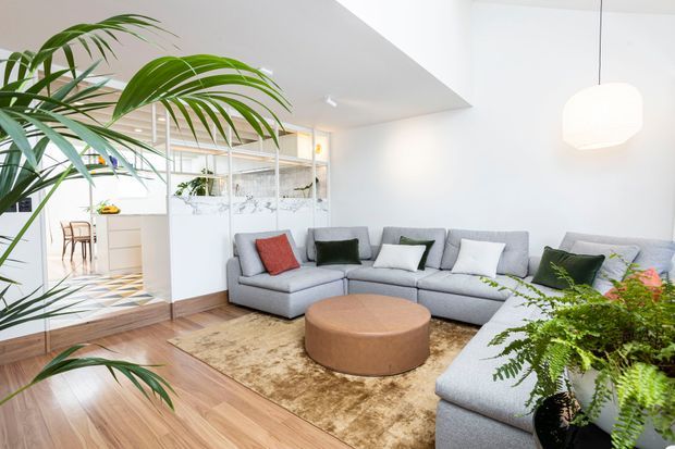 Bright contemporary living room with a large light-grey sectional sofa arranged around a round tan ottoman and textured rug. The room has warm timber flooring, white walls and ceiling, a suspended white pendant light, abundant indoor plants, and a glazed partition opening to a light-filled kitchen and dining area. A geometric tiled floor is visible in the adjoining space.