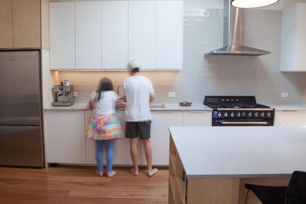 Bright contemporary kitchen with white flat-panel cabinetry, pale stone benchtops, a large island, stainless-steel refrigerator and rangehood, and a freestanding gas range. Warm timber flooring and under-cabinet lighting add contrast. Two people are using the kitchen, with a coffee machine and other small appliances visible along the rear counter.