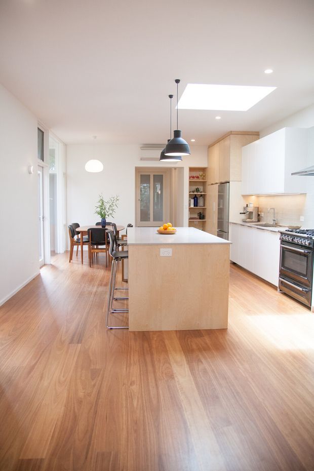 Bright open-plan kitchen and dining area with a large central island, light timber cabinetry and flooring, white cabinetry and benchtops, stainless-steel appliances, pendant lighting, skylight, and a small dining table beside tall windows.