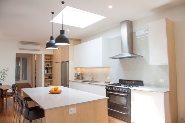 Bright contemporary kitchen with a large central island and integrated dining seating, white flat-panel cabinetry, pale timber island cladding, light benchtops, stainless-steel rangehood and freestanding cooker. Three black pendant lights hang beneath a large rectangular skylight, with open shelving and a glazed door visible at the rear.
