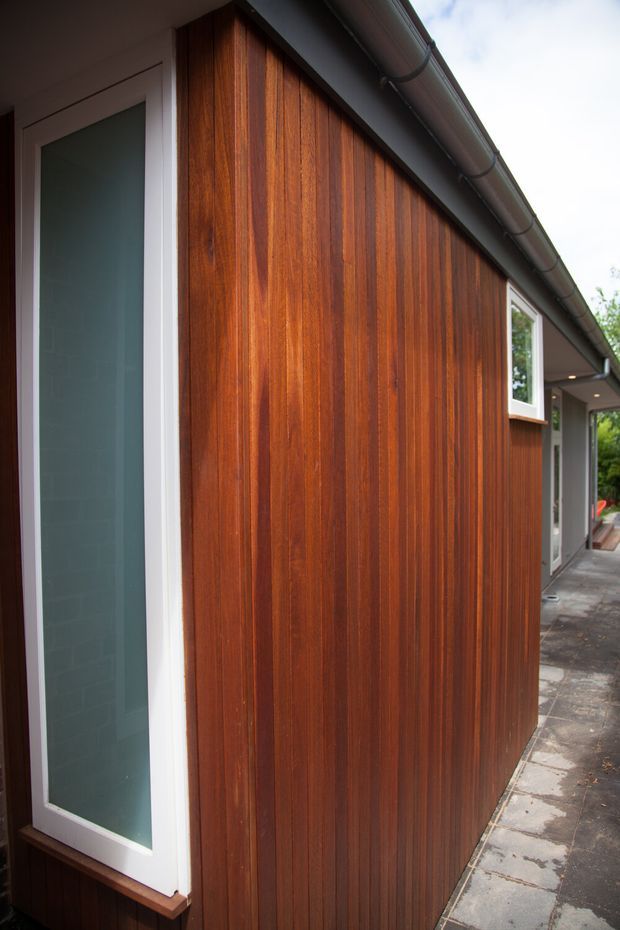 Close-up exterior view of a contemporary building extension with rich vertical timber cladding, white-framed glazed openings, a dark roof gutter, and a narrow concrete-paved path alongside the facade.