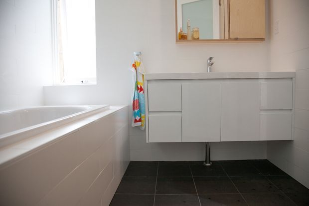 Bright, clean contemporary bathroom featuring a built-in white bathtub, wall-hung white vanity with integrated basin, mirrored timber-framed wall cabinet, white tiled walls, and dark floor tiles. A colourful patterned towel adds a small accent.
