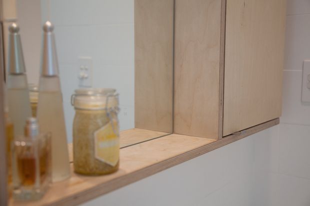 Close-up of a minimalist built-in timber wall cabinet and open shelf, with visible layered plywood edges. Toiletries and a glass jar sit on the shelf, while a mirror and light-coloured tiled wall are visible behind.