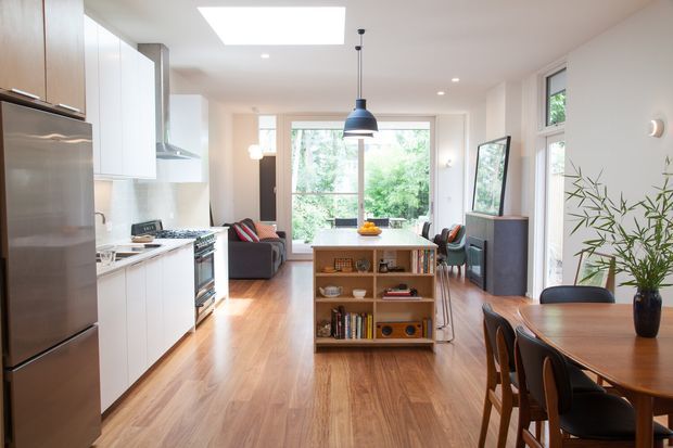 Bright open-plan interior combining a white kitchen, central timber island and dining area with a living space beyond. The room features warm timber flooring, white cabinetry, stainless-steel appliances, large windows, a skylight, and a black pendant light. A freestanding open shelving unit separates the kitchen from the living area.
