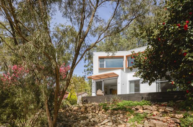 Exterior view of a contemporary two-storey residence set among mature trees and landscaped rock retaining walls. The white rendered facade has strong rectangular forms, large windows, dark horizontal cladding at the lower level, and a small covered entry or terrace with a translucent slatted roof.