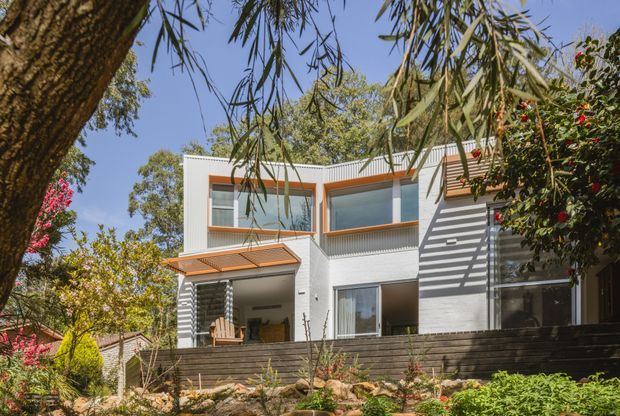 Exterior view of a contemporary two-storey residence set within a landscaped suburban garden. The facade combines white masonry walls, broad glazed openings with warm timber-toned frames, a corrugated metal roofline, and a covered entry or outdoor sitting area. Dark horizontal timber screening and a stepped garden terrace define the foreground, while mature trees frame the house.