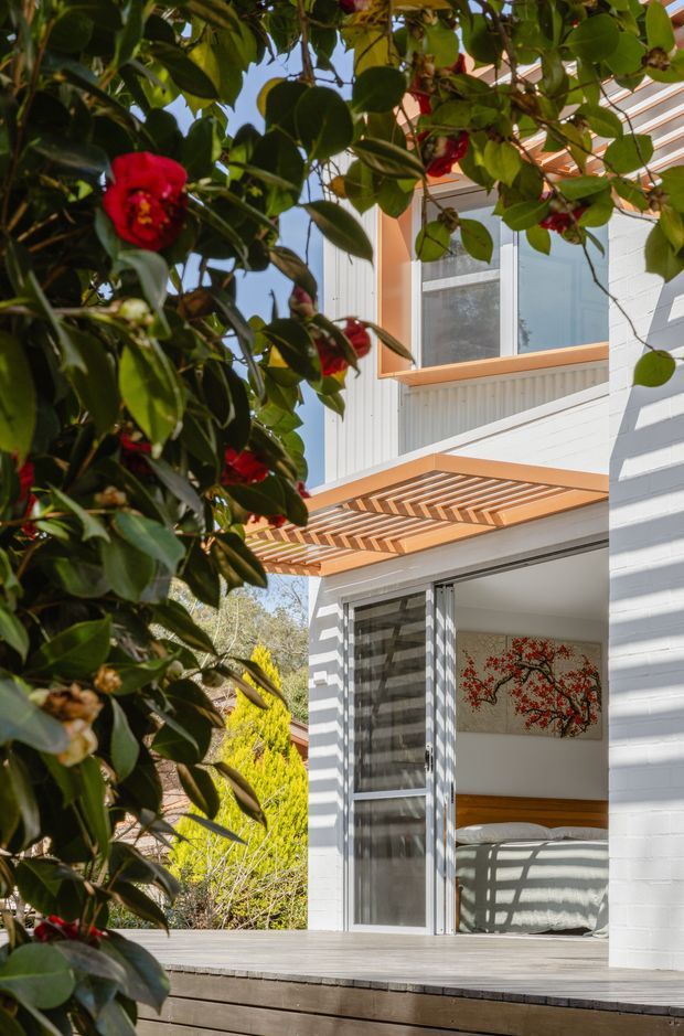 Vertical exterior view of a light-coloured, multi-level residential building partially framed by leafy flowering vegetation. White horizontal cladding, timber-framed windows, and a projecting timber pergola create a shaded outdoor threshold above a timber deck. Through the open ground-floor glazing, a bedroom with a bed and wall artwork is visible.
