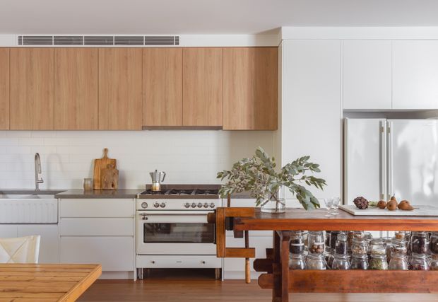 Bright contemporary kitchen with warm timber overhead cabinetry, white lower cabinets, a tiled backsplash, freestanding stainless-steel range, integrated refrigerator, and a substantial timber island or bar in the foreground. Natural branches and timber accessories add warmth to the otherwise clean, light interior.
