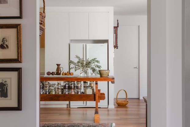 View through a wide doorway into a bright, compact kitchen with white full-height cabinetry and a white refrigerator, warm timber flooring, and a substantial timber-topped island or worktable. The island includes open shelving filled with jars, while ceramics, produce, framed artwork, and a basket add a curated traditional character. A hallway and closed white door are visible beyond.