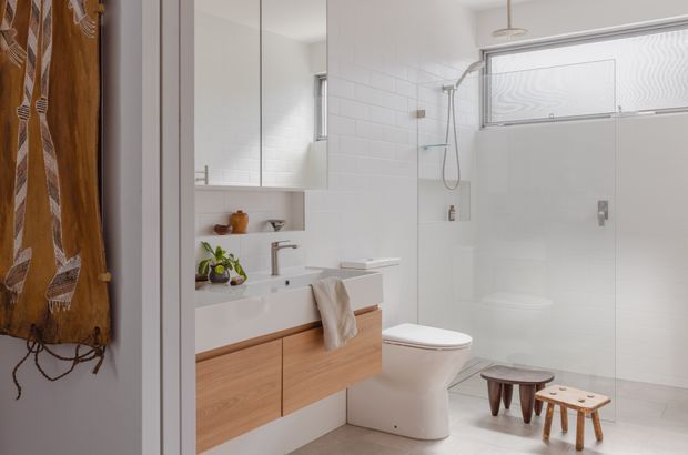 Bright contemporary bathroom viewed from an adjoining space, featuring a floating light-wood vanity with white basin, wall-mounted mirror cabinetry, white tiled walls, a frameless glass shower enclosure, wall-hung shower fittings, and a concealed-style toilet. Small wooden stools and a patterned textile add warmth to the otherwise clean, pale interior.