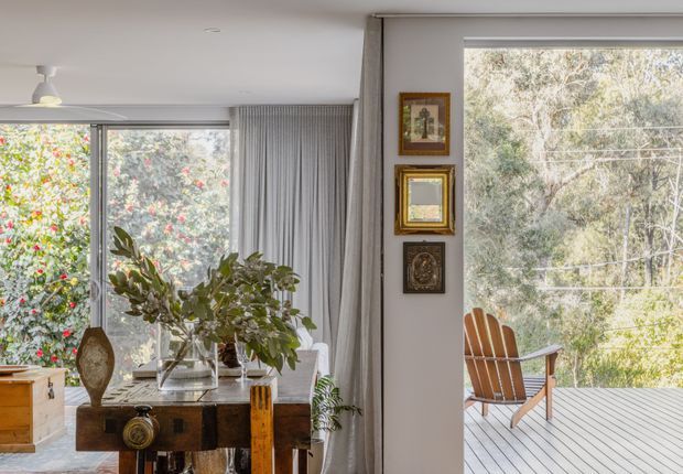 Bright mid-century-inspired living room opening onto a timber deck, with extensive glazing overlooking leafy vegetation. The room contains a vintage wooden console, decorative objects, indoor plants, framed artwork, curtains, and a hanging pendant light. An outdoor lounge chair is visible on the deck beyond the glazing.