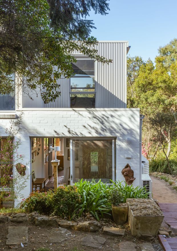Exterior view of a compact two-storey dwelling set within dense landscaped vegetation. The lower level has white-painted brick walls, broad glazed openings and a timber-framed entry, while the upper level is clad in vertically ribbed light-coloured metal or corrugated sheet material with a large horizontal window. A paved timber walkway, garden beds, rocks and mature trees surround the building.