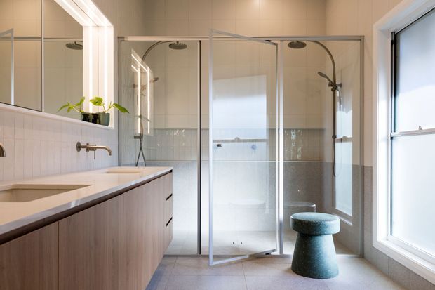 Bright contemporary bathroom with a long timber-look floating vanity, integrated basin, wall-mounted brushed metal tapware, large mirrored wall, and a glazed walk-in shower enclosure with dual shower fittings. Neutral tiled walls and floor are balanced by a sculptural dark teal stool and generous natural light from the window.