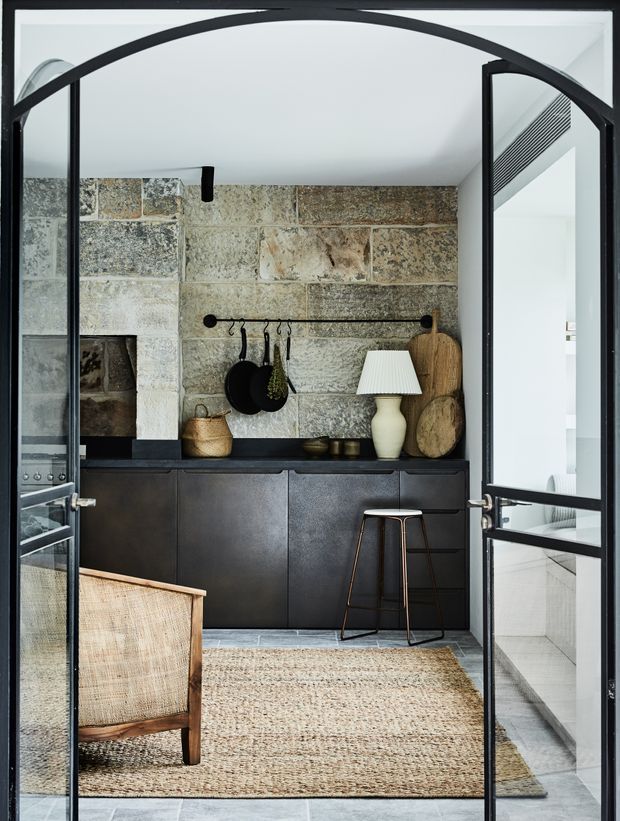 A compact, richly textured kitchen or kitchenette viewed through black-framed glazed doors. The room features a large reclaimed-looking stone wall and backsplash, dark built-in cabinetry, dark benchtops, open rail storage with cookware, a woven rug, a cane chair, and a small bar stool. A bright adjacent space with pale tiled surfaces is visible through the opening on the right. The overall design combines rustic natural materials with refined contemporary detailing.