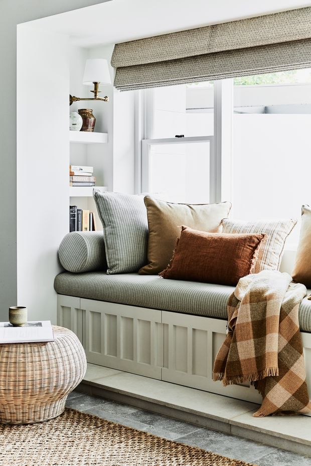 Bright, inviting window-seat nook with a built-in upholstered bench beneath a large window. White painted panelled joinery and shelving frame the seat, with woven roller blinds, layered neutral and rust-coloured cushions, a textured throw, a wicker coffee table, and a natural woven rug over grey stone-look flooring.