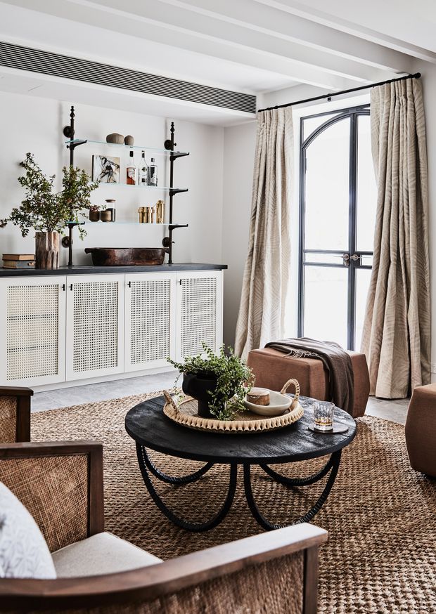 A refined living room with a neutral palette, featuring a black circular coffee table, woven rug, cane-front built-in cabinetry, open display shelving, upholstered lounge chairs, and full-height textured curtains. The room has white exposed ceiling beams, a dark-framed arched window or glazed door, and layered natural materials for a warm, sophisticated atmosphere.