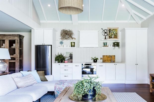 Bright open-plan living and kitchen interior with a white built-in kitchen, integrated appliances, tall cabinetry, vertical wall panelling, and a vaulted ceiling with exposed white-painted beams. A pale sectional sofa, timber coffee table, woven pendant light, and neutral textiles create a relaxed coastal feel. A rustic timber cabinet is visible at left.