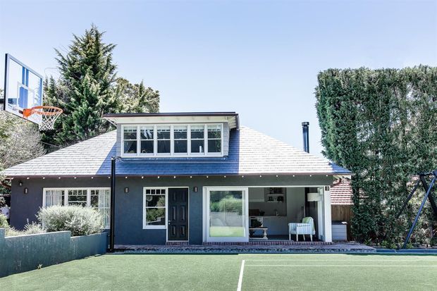 Front exterior view of a suburban house with a dark painted lower facade, pale shingle-clad pitched roof, prominent dormer window, covered patio opening, and broad synthetic lawn or sports court in the foreground. A basketball hoop and children's swing set are visible around the landscaped residential setting.
