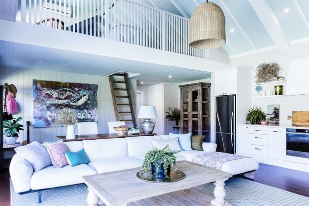 Bright open-plan living room with a large white sectional sofa, pale timber coffee table, built-in white kitchen cabinetry, and a loft-style mezzanine with white vertical balustrades. The room features a coastal palette, exposed light-painted ceiling boards, a large woven pendant light, artwork, plants, and a ladder leading to the upper level.