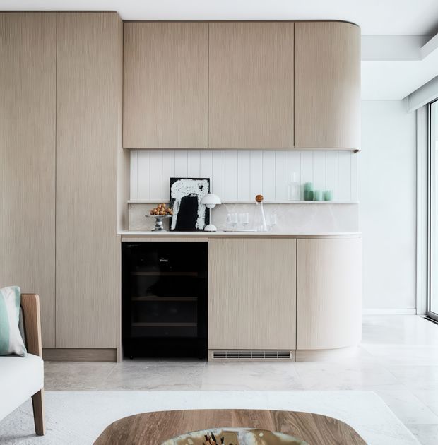 A compact, refined kitchen integrated into a bright open-plan interior. Floor-to-ceiling light timber cabinetry features fine vertical grain, rounded corners, and concealed storage, paired with a pale stone benchtop and white vertically panelled splashback. A black built-in oven provides contrast, while full-height glazing at right brings in abundant natural light. Part of a pale upholstered chair and a rounded timber coffee table are visible in the foreground.
