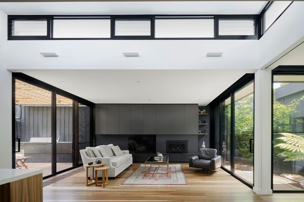Bright contemporary living room with a double-height ceiling and clerestory windows. The space features extensive black-framed glazing, warm timber flooring, a dark built-in media and fireplace wall with open shelving, and a seating arrangement of a light sofa, armchair, coffee table, and patterned rug. A partially visible kitchen island appears in the foreground, while a covered outdoor area and greenery are visible through the glazing.