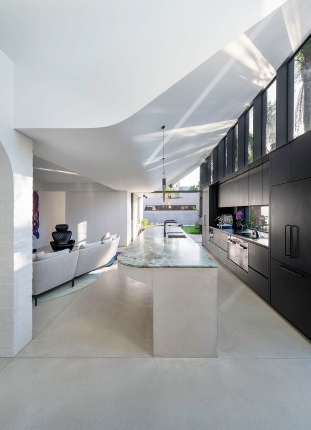 Bright open-plan contemporary kitchen and living area with a sculptural curved white ceiling, full-height dark-framed glazing, dark built-in kitchen cabinetry, and a large pale stone island with a softly veined surface. A light sofa and black lounge chair sit beyond the island, while daylight enters from the glazed side and rear courtyard.