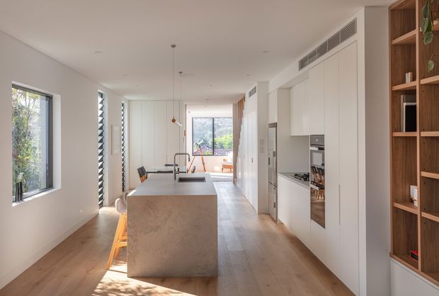 Bright contemporary open-plan kitchen viewed along a long, narrow interior. A large stone island with an undermount sink and dark tap anchors the room, with timber flooring, white full-height cabinetry, integrated appliances, built-in timber shelving, pendant lights, and extensive windows providing natural light. A dining or living area is visible beyond the kitchen.