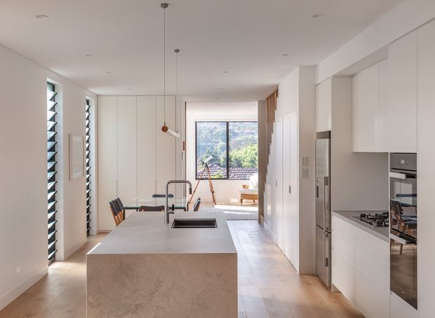 A bright, narrow contemporary kitchen viewed along a long open-plan layout. The room features a large pale stone island with an integrated sink and dark tapware, white floor-to-ceiling cabinetry, a built-in oven and cooktop, pale timber flooring, slim pendant lights, louvred windows on the left, and glazing at the far end opening toward a leafy view and small outdoor seating area. The design is clean-lined and minimalist with abundant natural light.