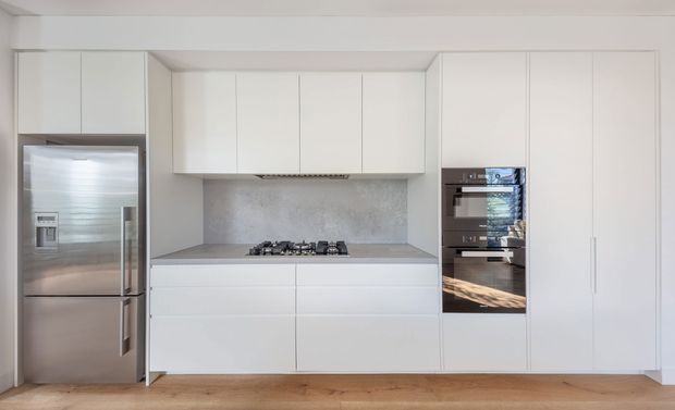 A clean, contemporary kitchen with a wall of white flat-panel cabinetry, integrated appliances, a stainless-steel refrigerator, gas cooktop, and built-in black-glass ovens. The cabinetry surrounds a recessed cooking zone with a textured light-grey splashback, while warm timber flooring adds contrast.