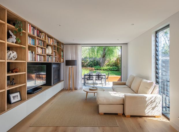 Bright contemporary living room with extensive built-in timber shelving and cabinetry along the left wall, incorporating a television and fireplace. A large cream sectional sofa faces the joinery, with a neutral area rug and low round coffee table. Full-height glazing and a glass door open to a furnished timber deck and leafy garden, while a louvred window on the right brings in additional light. The room has a clean, warm, indoor-outdoor character.