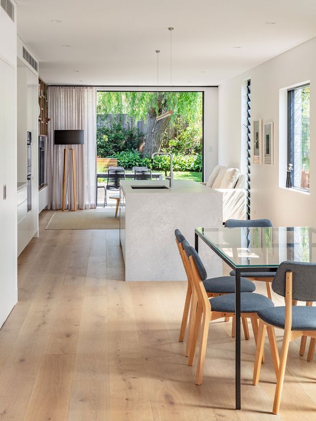 A bright, open-plan contemporary kitchen and dining area with light timber flooring, white built-in cabinetry, and a large pale stone island with an integrated sink and gooseneck tap. A glass dining table with timber-framed upholstered chairs occupies the foreground. Full-height glazing opens toward a leafy garden, while sheer curtains, a streamlined living area, and narrow high-level ventilation windows contribute to an airy, modern interior.